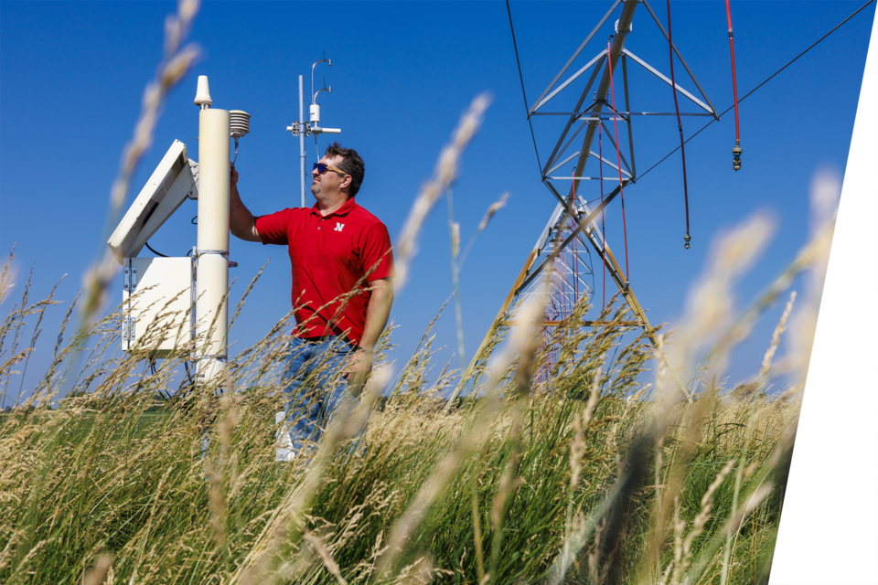 A man in a red Nebraska shirt inspects a solar panel connection to his pivots in a wheat field.
